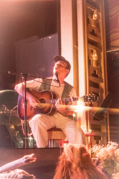 Mac Demarco onstage singing and playing guitar at Webster Hall. He is wearing all white despite a green vest and dark hat, and he is looking upwards. The back of fans