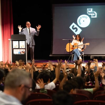 A speaker on stage with an artist seated beside him holding an acoustic guitar, the Sounds of Saving logo projected behind, and a full auditorium of students with hands raised