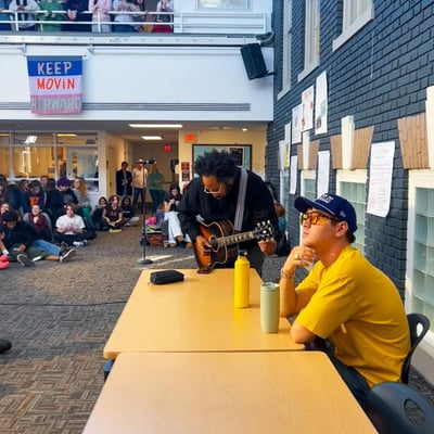 An artist leaning over a table playing an acoustic guitar for a student, with more students seated on the floor and watching from a balcony above