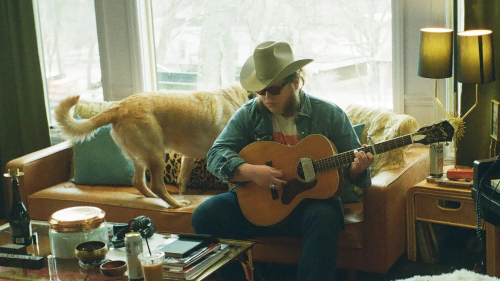 A musician playing guitar in a living room, a dog walking past