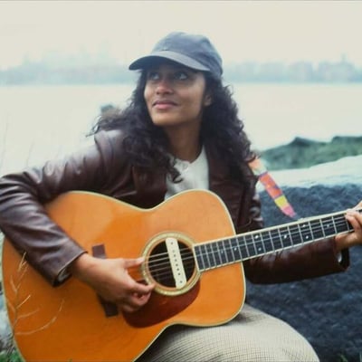 An artist playing acoustic guitar on a shoreline during a filmed session